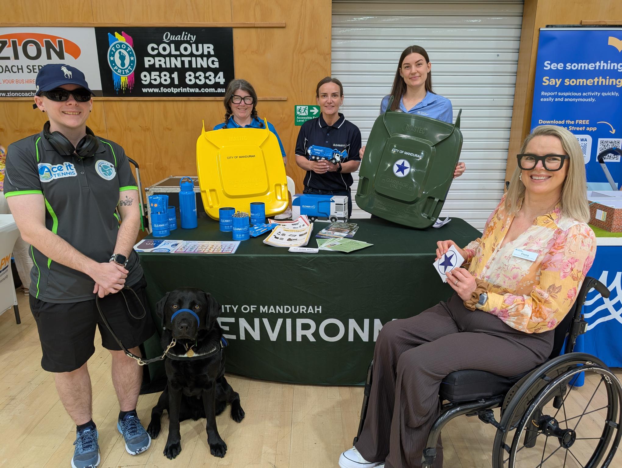 A group of five people and a black guide dog pose around a City of Mandurah community information table showcasing tactile waste bin lids designed for accessibility.
